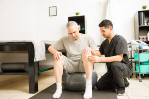 Physical therapist assisting mature male patient sitting on exercise ball in clinic