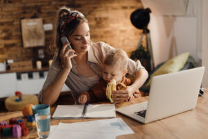 Single mother feeding her son with a banana while communicating on mobile phone and working at home